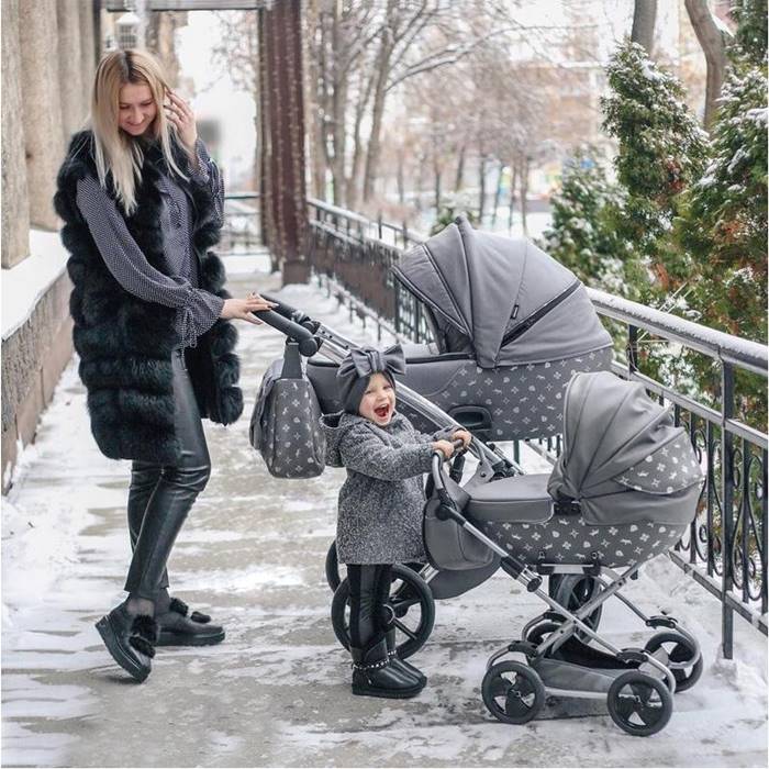 Mama i córka podczas zimowego spaceru – każda z własnym wózkiem. Mama prowadzi dziecięcy wózek, a dziewczynka swoją mini wersję dla lalek. Obie ubrane ciepło, w tle śnieżna sceneria.